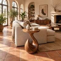 Circular side table on a teardrop pedestal with a swirling neck in a bright living room with arched windows and plants