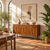 Three door sideboard with carved face reliefs and tapered angled legs in a bright dining room with arched windows and plants