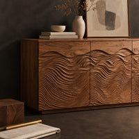 Corner detail of low sideboard with three doors carved in flowing waves on a plinth base beside wood, brass and linen samples
