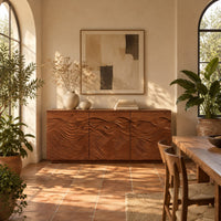 Low sideboard with three doors carved in flowing waves on a plinth base in a bright dining room with arched windows and plants
