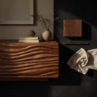 Corner detail of media cabinet with wave carved doors around an open niche beside wood, brass and linen samples