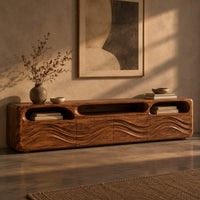 Corner detail of low tv stand with carved wave relief on the front and open top cubbies beside wood, brass and linen samples