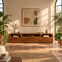 Low tv stand with carved wave relief on the front and open top cubbies in a bright living room with arched windows and plants