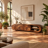 Tv stand with three wave carved fronts and an open looped end shelf in a bright living room with arched windows and plants