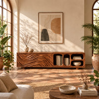 Low tv stand with wave carved fronts and open pill shaped cubbies in a bright living room with arched windows and plants