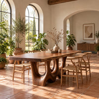 Dining table with a natural edge slab top and two sculpted arch legs in a bright dining room with arched windows and plants