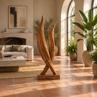 Twin feather sculpture with carved veins crossing over a solid wood plinth in a bright living room with arched windows and plants