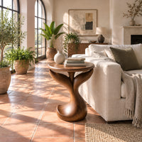 Pedestal side table with a round top, twin fins and a curled foot in a bright living room with arched windows and plants