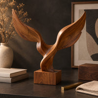 Corner detail of twin wing sculpture with swept tips rising from a square wooden block beside wood, brass and linen samples
