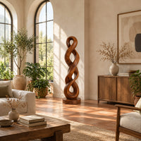 Twisted wood sculpture of two intertwined loops with a flat disc foot in a bright living room with arched windows and plants