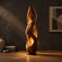 Tall twisted wooden floor lamp with open cutouts that glow warm amber in a dark studio against a charcoal plaster wall