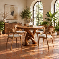 Circular wooden dining table with a twisting braided pedestal on a disc base in a bright dining room with arched windows and plants