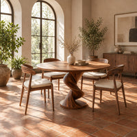 Round dining table with a twisting spiral pedestal on a circular foot in a bright dining room with arched windows and plants