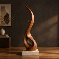 Tall twisting flame shaped wood sculpture with an open loop near the base in a dark studio against a charcoal plaster wall