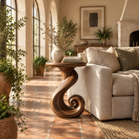 Round side table on a ribbon pedestal that ends in a spiral curl in a bright living room with arched windows and plants