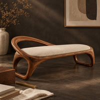 Corner detail of bench with a padded seat and an arched back rail on open loop legs beside wood, brass and linen samples