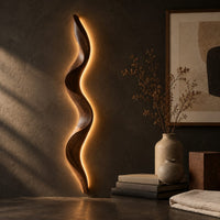 Corner detail of tall wavy wall sconce that glows softly along its back edge beside wood, brass and linen samples