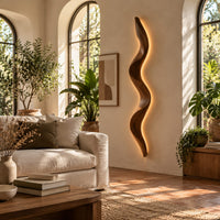 Tall wavy wall sconce that glows softly along its back edge in a bright living room with arched windows and plants