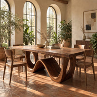 Rectangular dining table with a wave shaped base and open arch ends in a bright dining room with arched windows and plants
