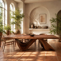 Rectangular dining table with a thick slab top on two ribbon like wave legs in a bright dining room with arched windows and plants