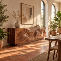 Long sideboard with deep wave carving across the full front in a bright dining room with arched windows and plants