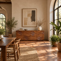 Wave carved wooden sideboard with four relief panels on a plinth base in a bright dining room with arched windows and plants