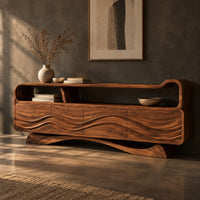 Corner detail of tv stand with wave carved panels, an open top shelf and an arched base beside wood, brass and linen samples