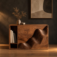 Boxy side table with a carved wave front and an open book slot in a dark studio against a charcoal plaster wall
