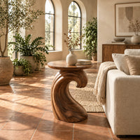 Side table with a wave shaped pedestal and a circular top in a bright living room with arched windows and plants