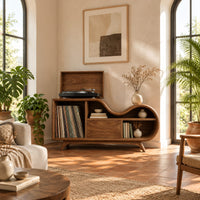 Low wooden record player stand with a wavy top and open record bay in a bright living room with arched windows and plants