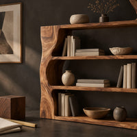 Corner detail of bookshelf with wavy wood sides, open shelves and a top ledge beside wood, brass and linen samples