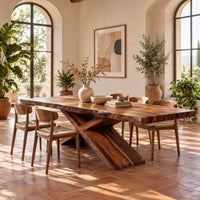 Dining table with crossed legs and a thick rectangular top in a bright dining room with arched windows and plants