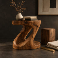 Corner detail of z shaped side table with a carved diagonal opening and wide base beside wood, brass and linen samples